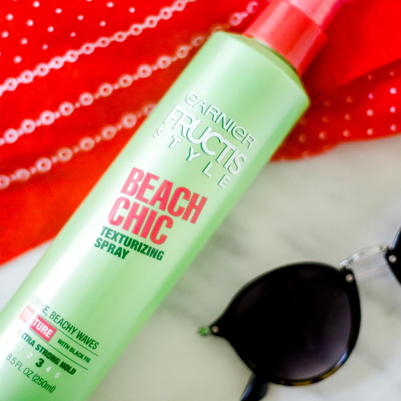 Garnier Fructis Style Beach Chic Texturizing Spray on a red handkerchief with white patterns on white marble next to black sunglasses.